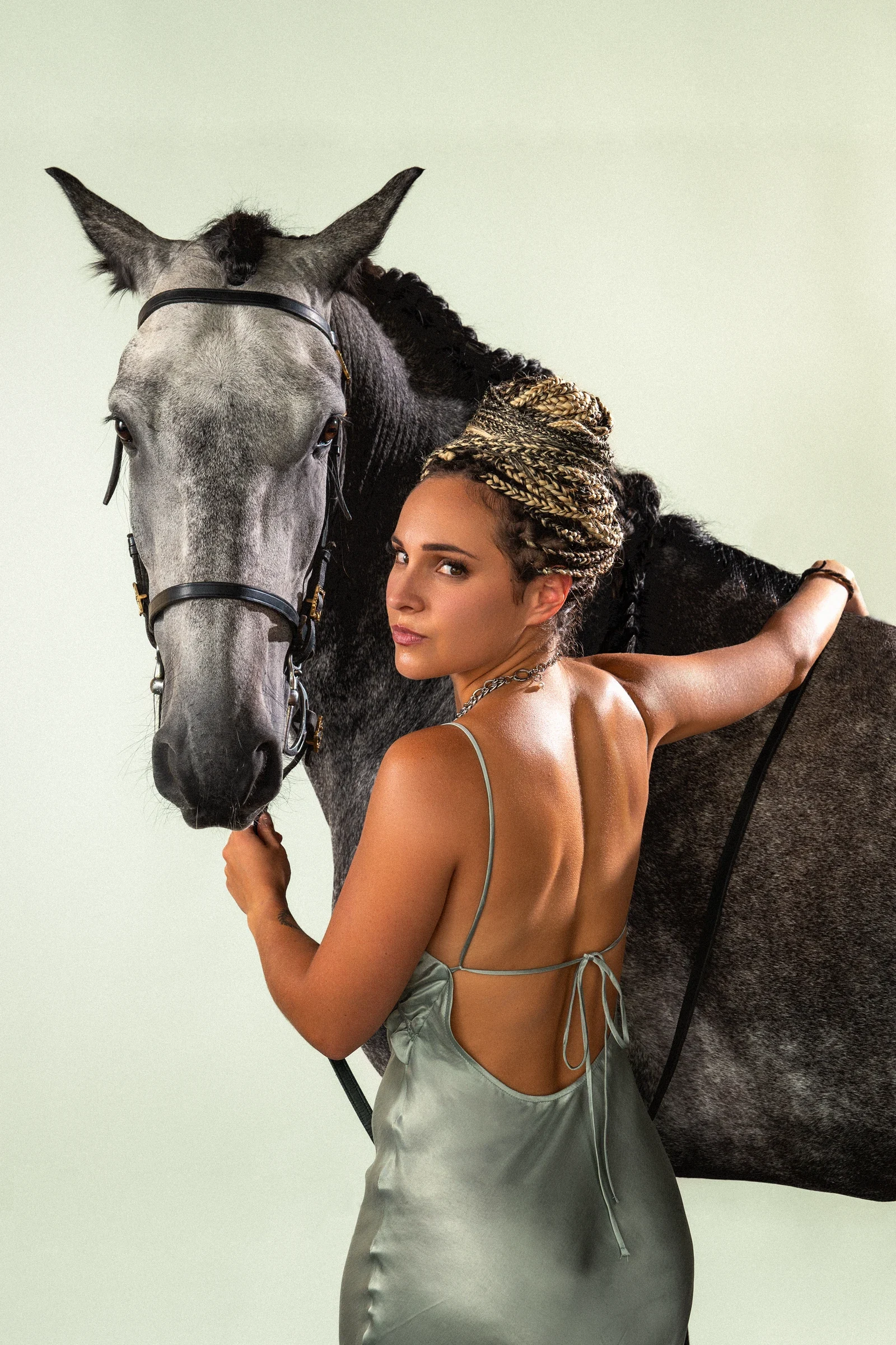 Photographie livrée Soft Horse, modèle de dos en robe satinée gris vert tenant un cheval gris contre elle