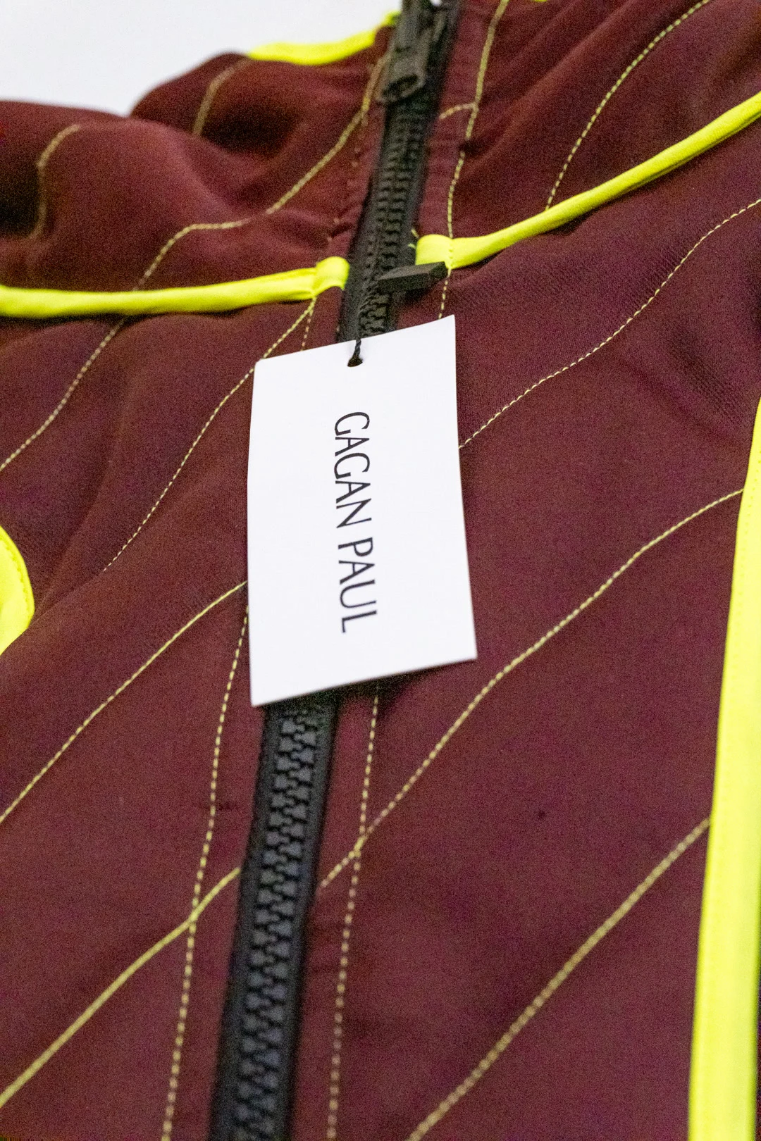 Gros plan sur une veste bordeaux Gagan Paul à surpiqûres jaunes avec étiquette blanche suspendue à la fermeture