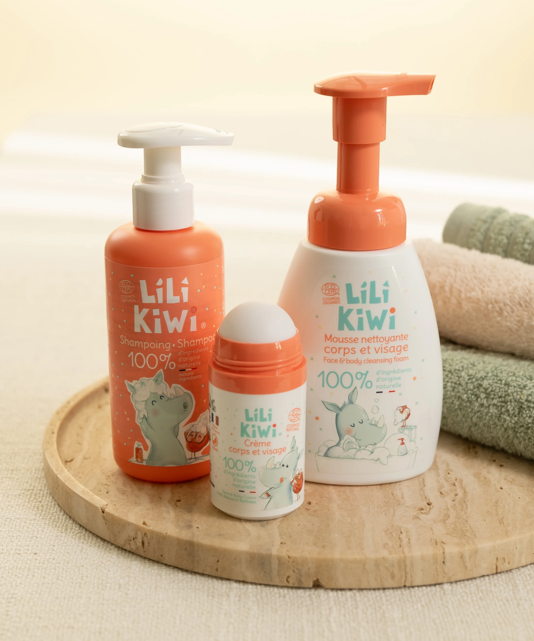 Trois produits Lili Kiwi orange et blanc réunis sur un plateau minéral avec du linge de bain