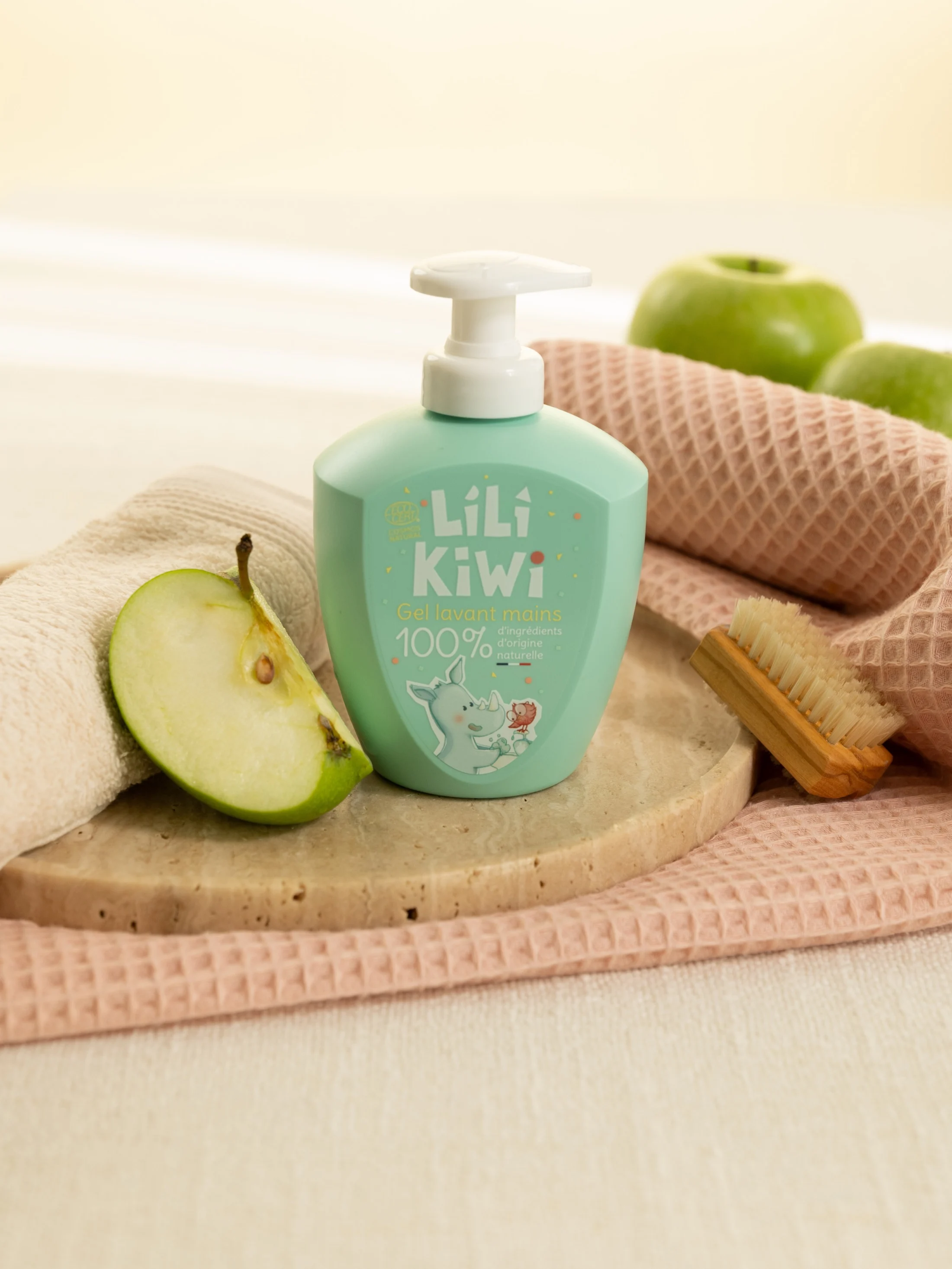 Gel lavant Lili Kiwi vert d’eau mis en scène avec une poire, du linge rose et une brosse en bois