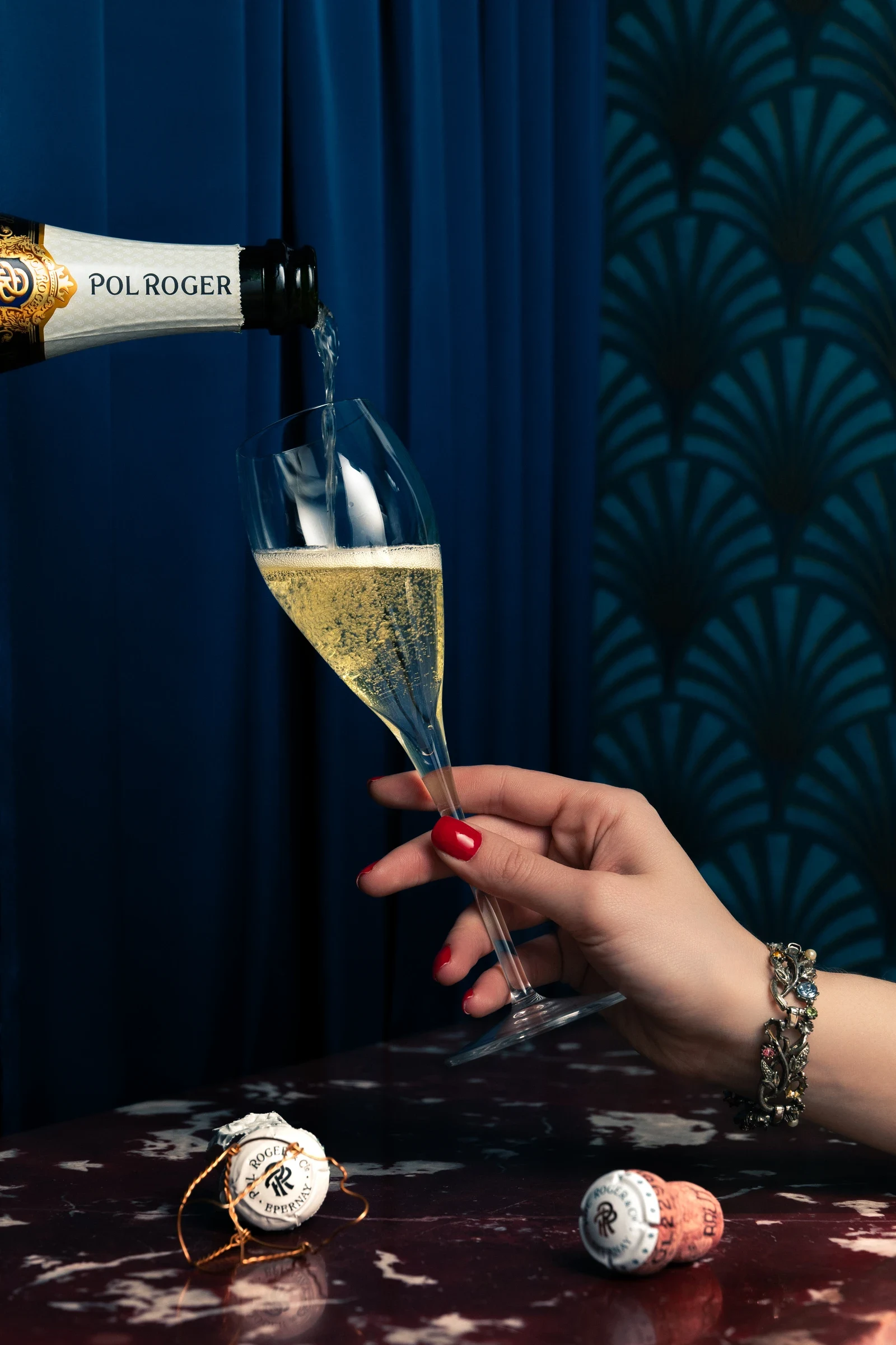 Champagne Pol Roger versé dans une flûte tenue à la main, devant un rideau bleu et une table de marbre