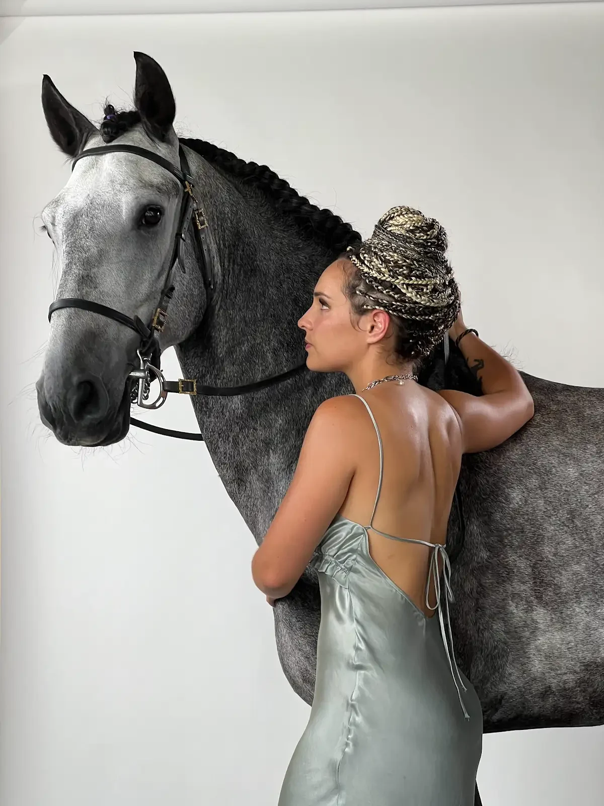 Portrait d’une femme en robe satinée gris vert auprès d’un cheval gris natté en studio