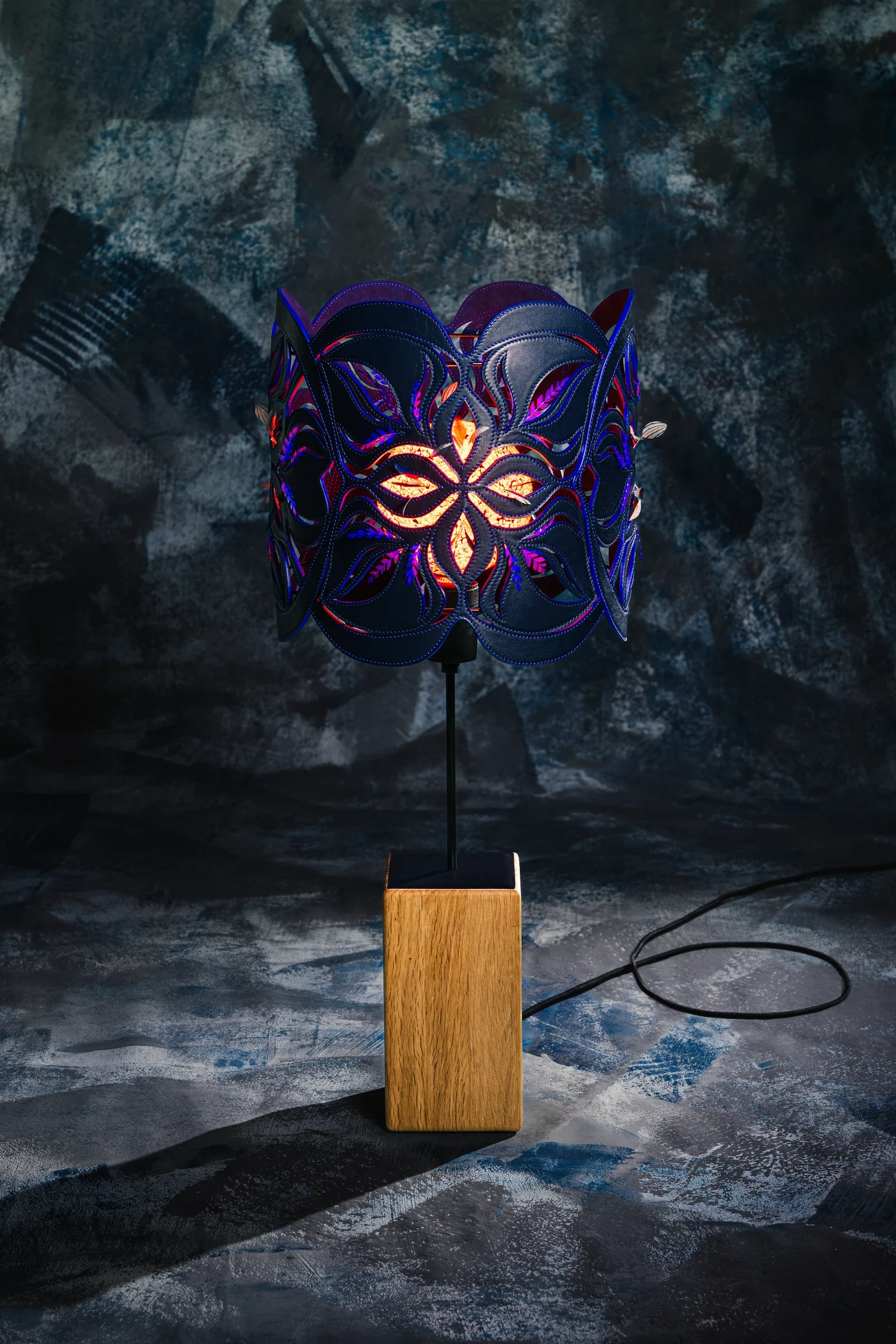 Lampe Prémisse allumée, dont la lumière chaude traverse un abat-jour ajouré bleu et violet, sur un socle de bois