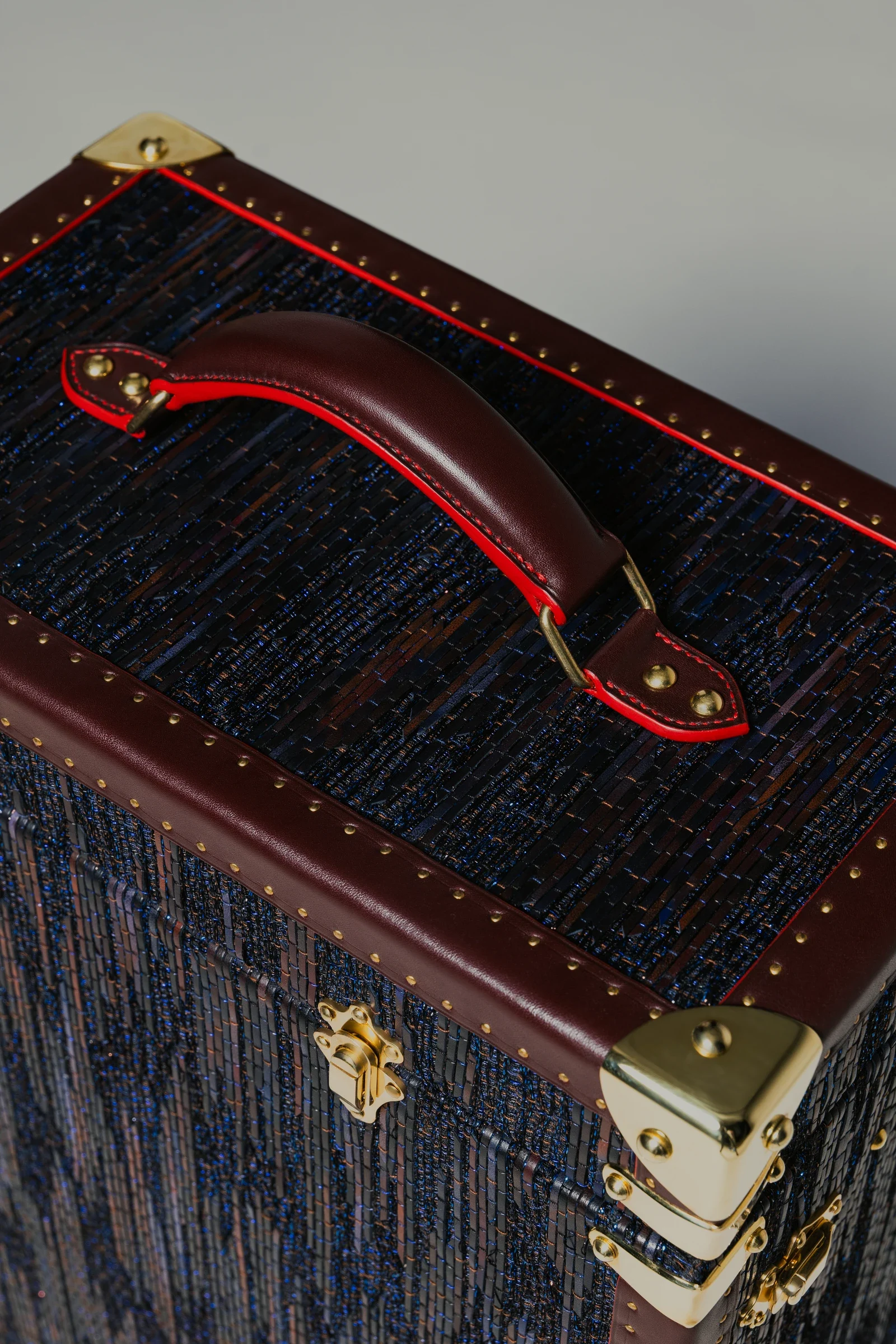 Détail de la poignée de cuir bordeaux, des coutures rouges et des ferrures dorées de la malle Prémisse